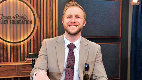 A smiling man in a suit sits in front of a wooden backdrop with the logo "Texas Public Policy Foundation"