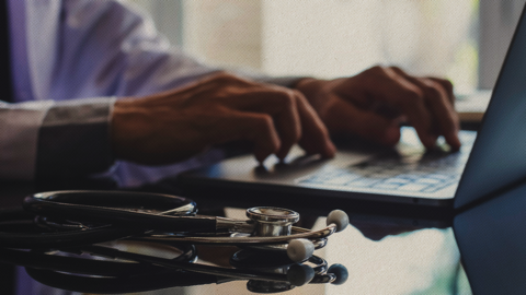 A doctor’s hands type on a laptop, with a stethoscope on the desk, representing telehealth and digital healthcare services.