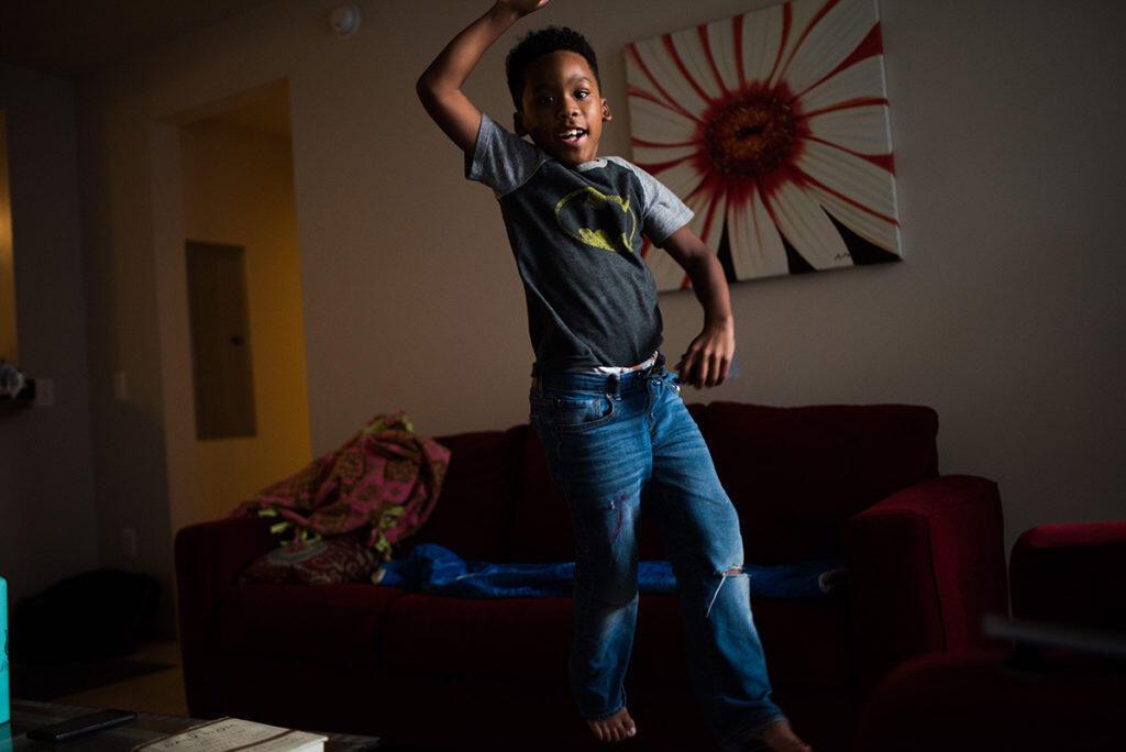 A boy in a batman shirt jumps in a living room